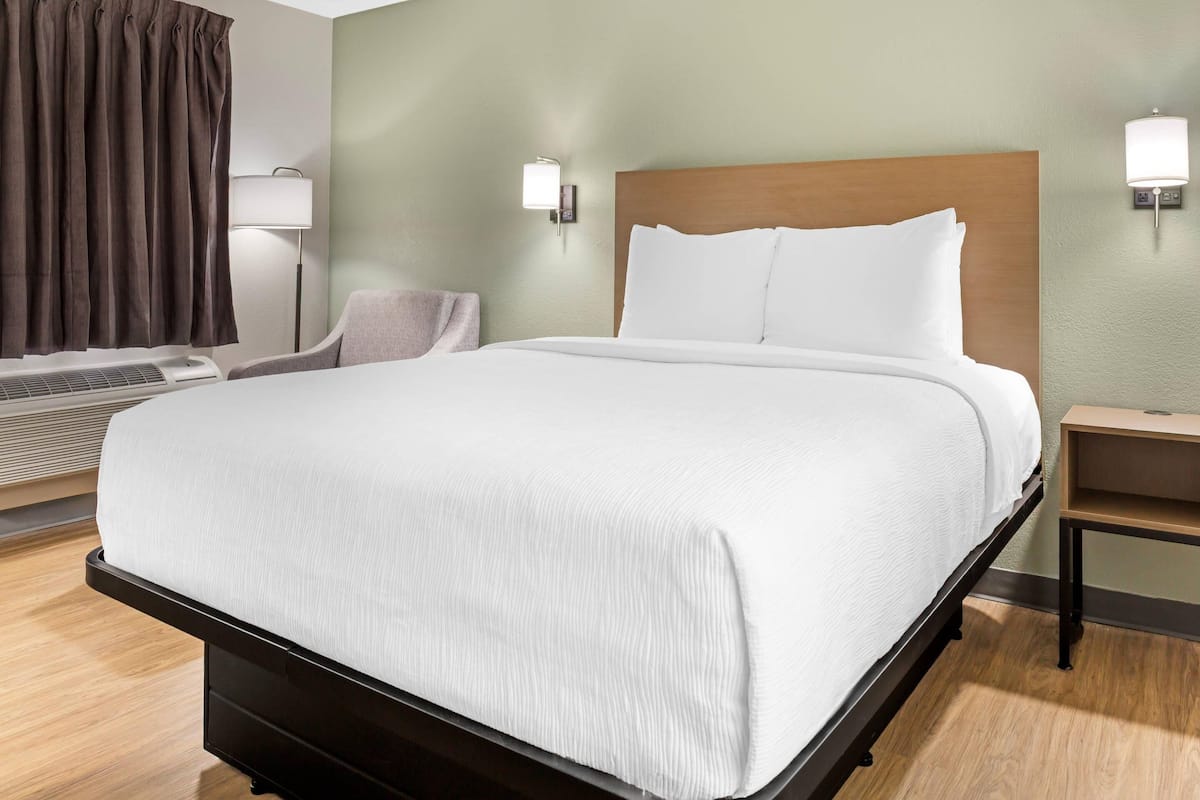 A neatly made bed with a wooden headboard, a bedside table, a lamp, and a chair in a hotel room.