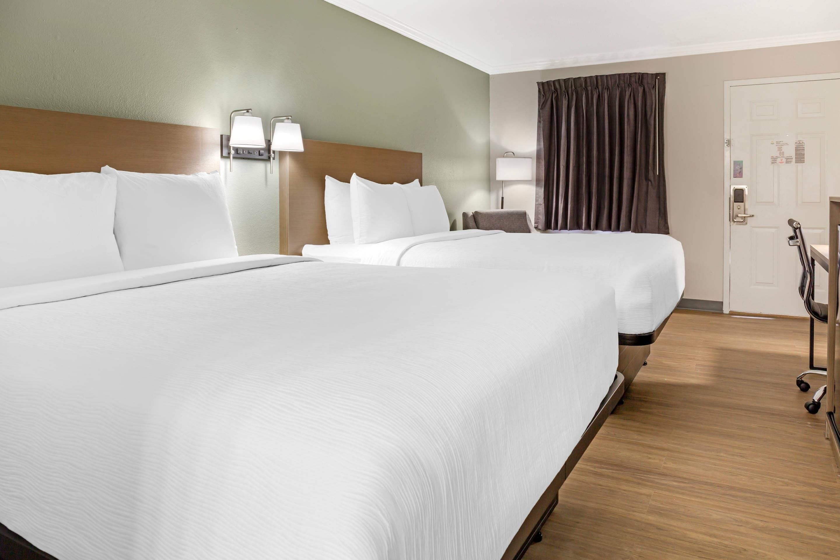 A hotel room with two beds, a wooden headboard, a nightstand with a lamp, a window with curtains, and a door with a lock.
