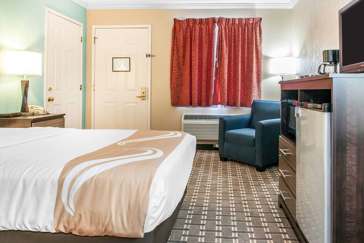 A hotel room with a bed, a desk with a lamp, a chair, a television, and a refrigerator.