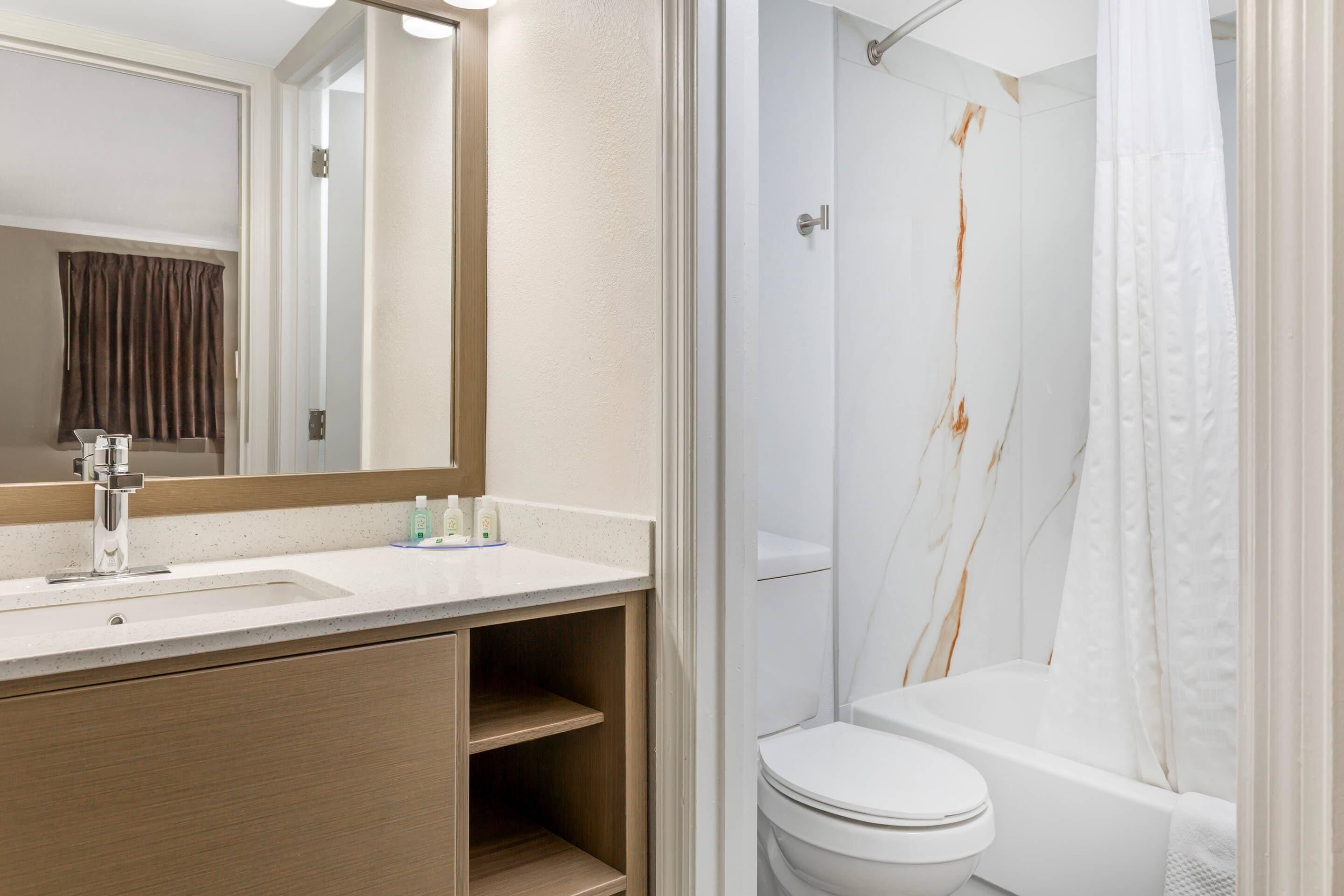 Une salle de bain avec un lavabo, un miroir et des toilettes blancs, une douche avec un rideau blanc et un mur en marbre.