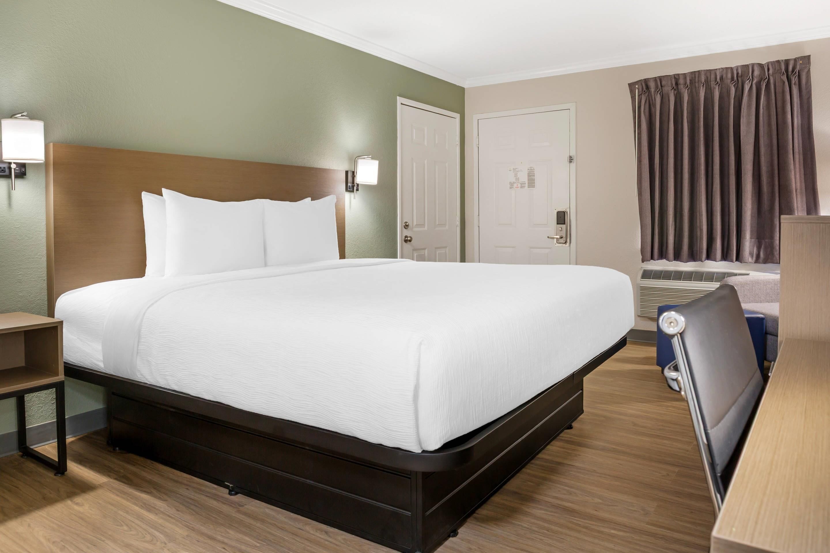 A hotel room with a large bed, a nightstand, a desk, a chair, and a window with curtains.