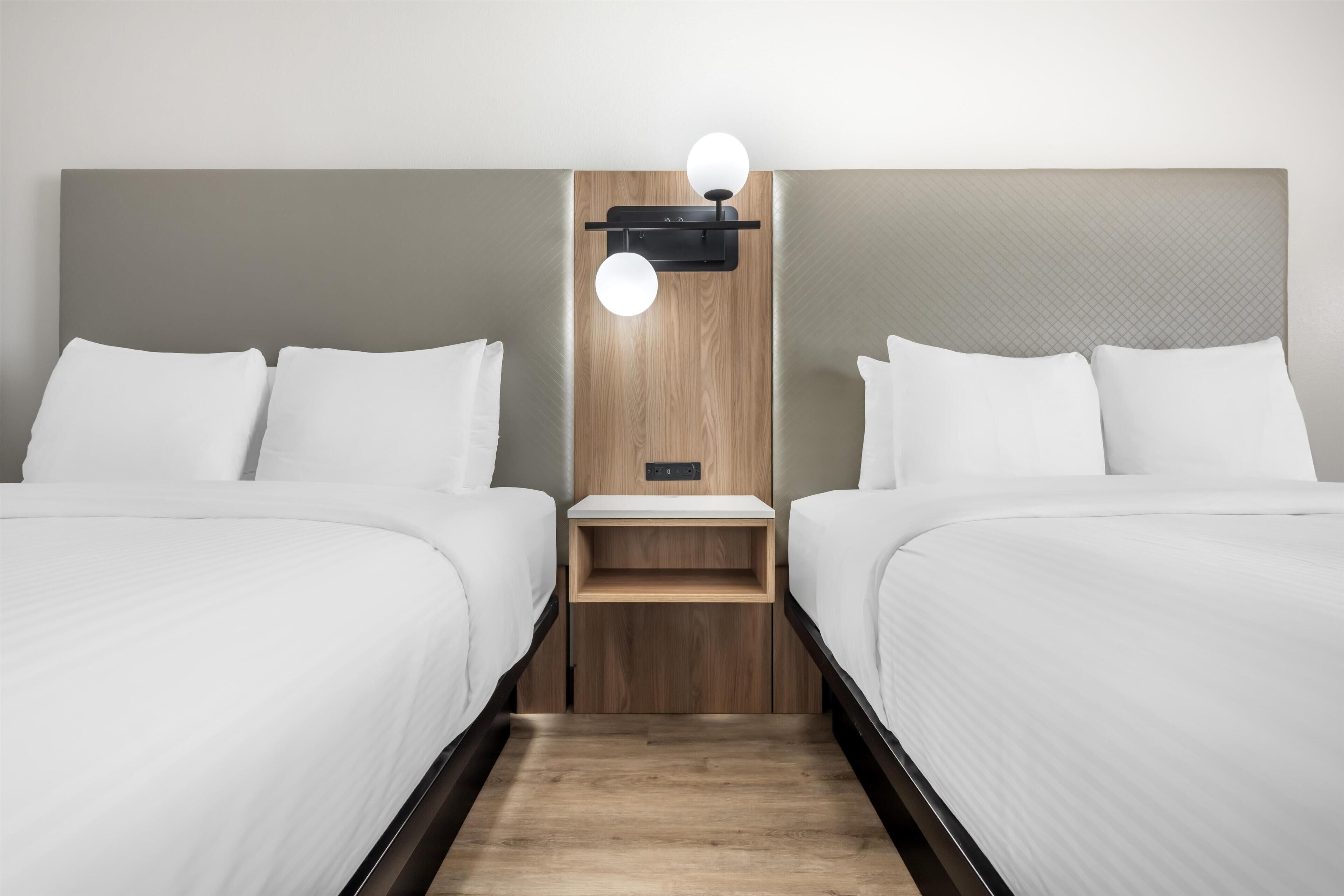 Habitación de hotel con dos camas, cabecera de madera, mesita de noche y lámpara de pared.
