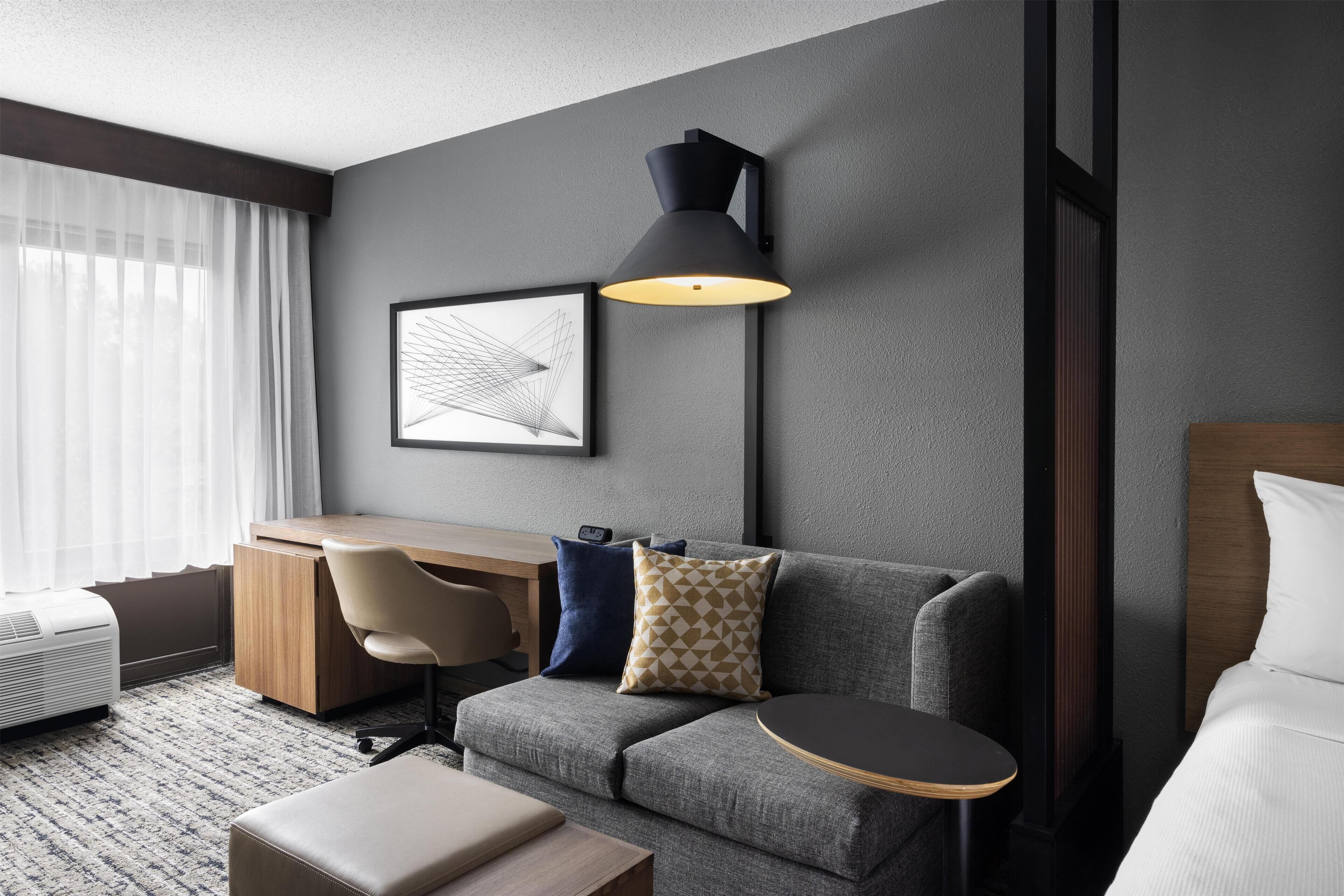 Una habitación de hotel moderna con un sofá gris, un escritorio de madera, una lámpara negra y un cuadro en la pared.