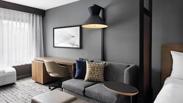 Quarto de hotel moderno com sofá cinzento, escrivaninha de madeira, luminária preta e quadro emoldurado na parede.