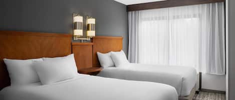 A hotel room with two beds, a wooden headboard, a nightstand, and a window with sheer curtains.