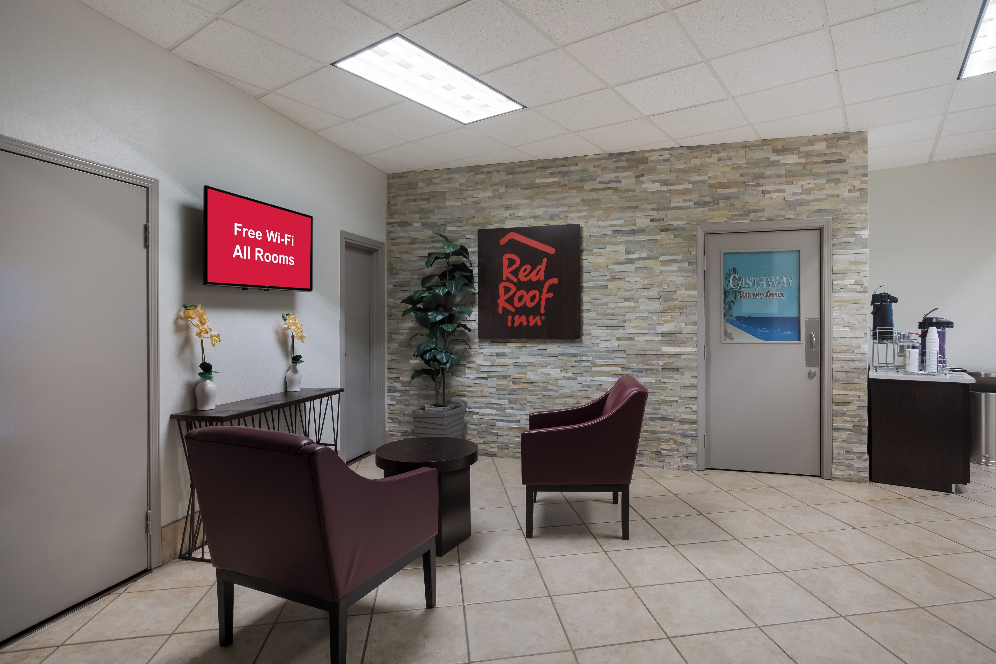 Zona de recepción con un televisor de pantalla plana mostrando 'Wi-Fi gratis en todas las habitaciones', cartel de Red Roof Inn, área de bar y un espacio de descanso con dos sillones.