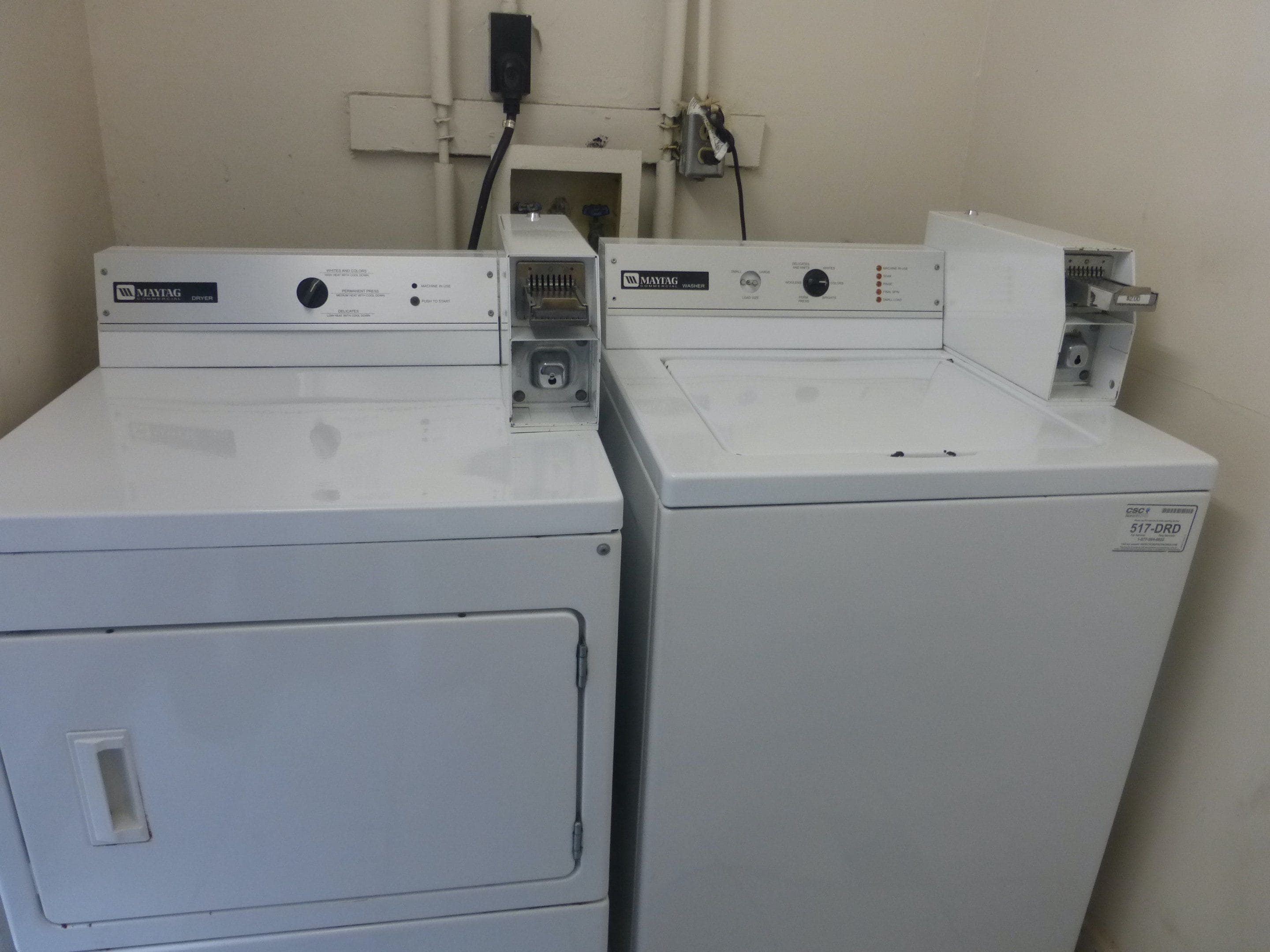 Dos lavadoras blancas Maytag en un cuarto de servicio con pared blanca y un enchufe eléctrico visible.