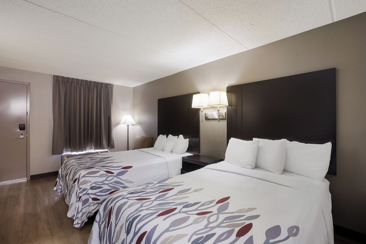 Habitación de hotel con dos camas, un cabecero grande y oscuro, una ventana con cortinas y una lámpara en la mesita de noche.