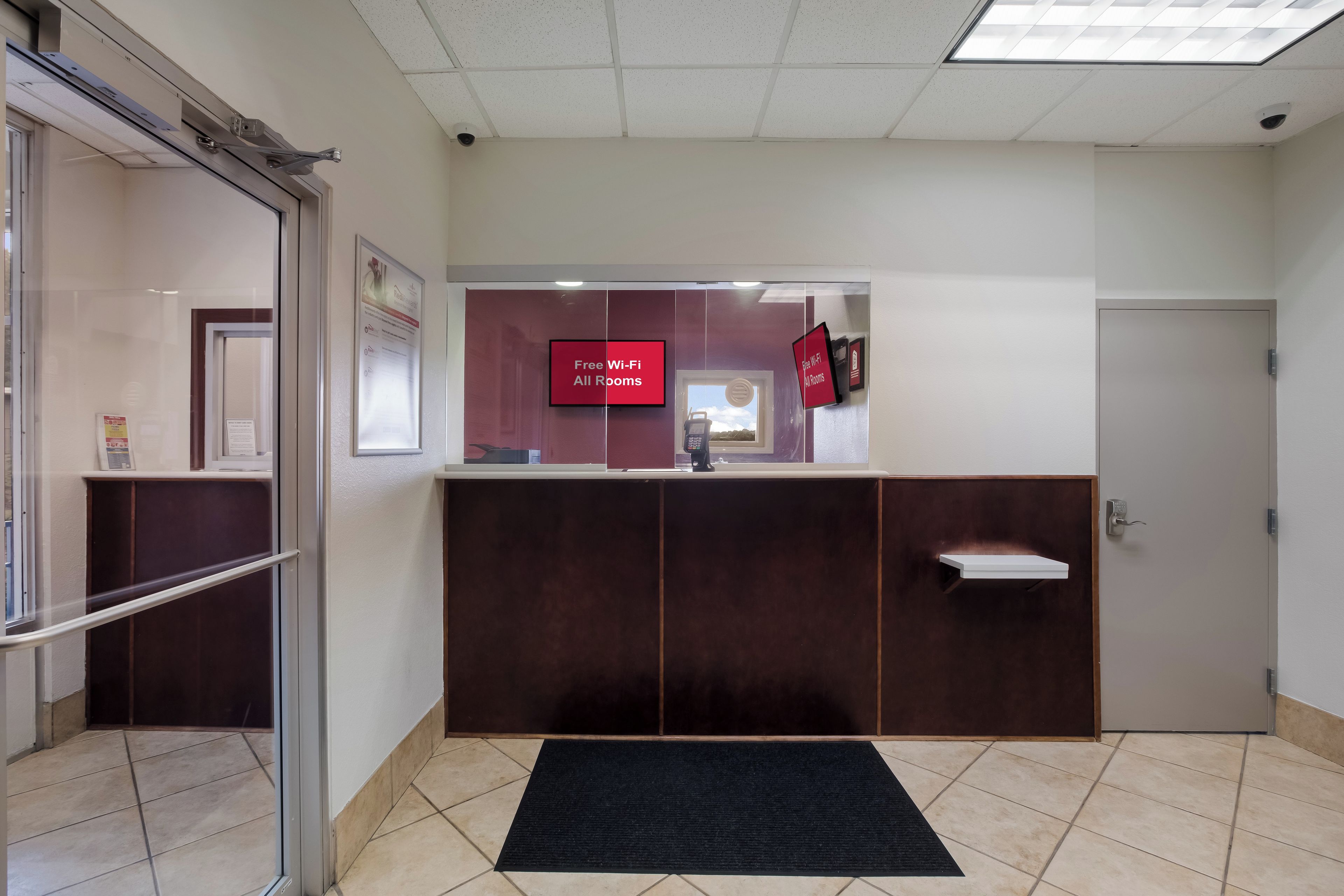A reception area with a glass door, a reception desk, a digital screen displaying "Free Wi-Fi All Rooms," and a small window.