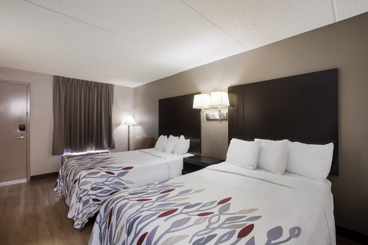 A hotel room with two beds, a large dark headboard, a window with curtains, and a lamp on the nightstand.