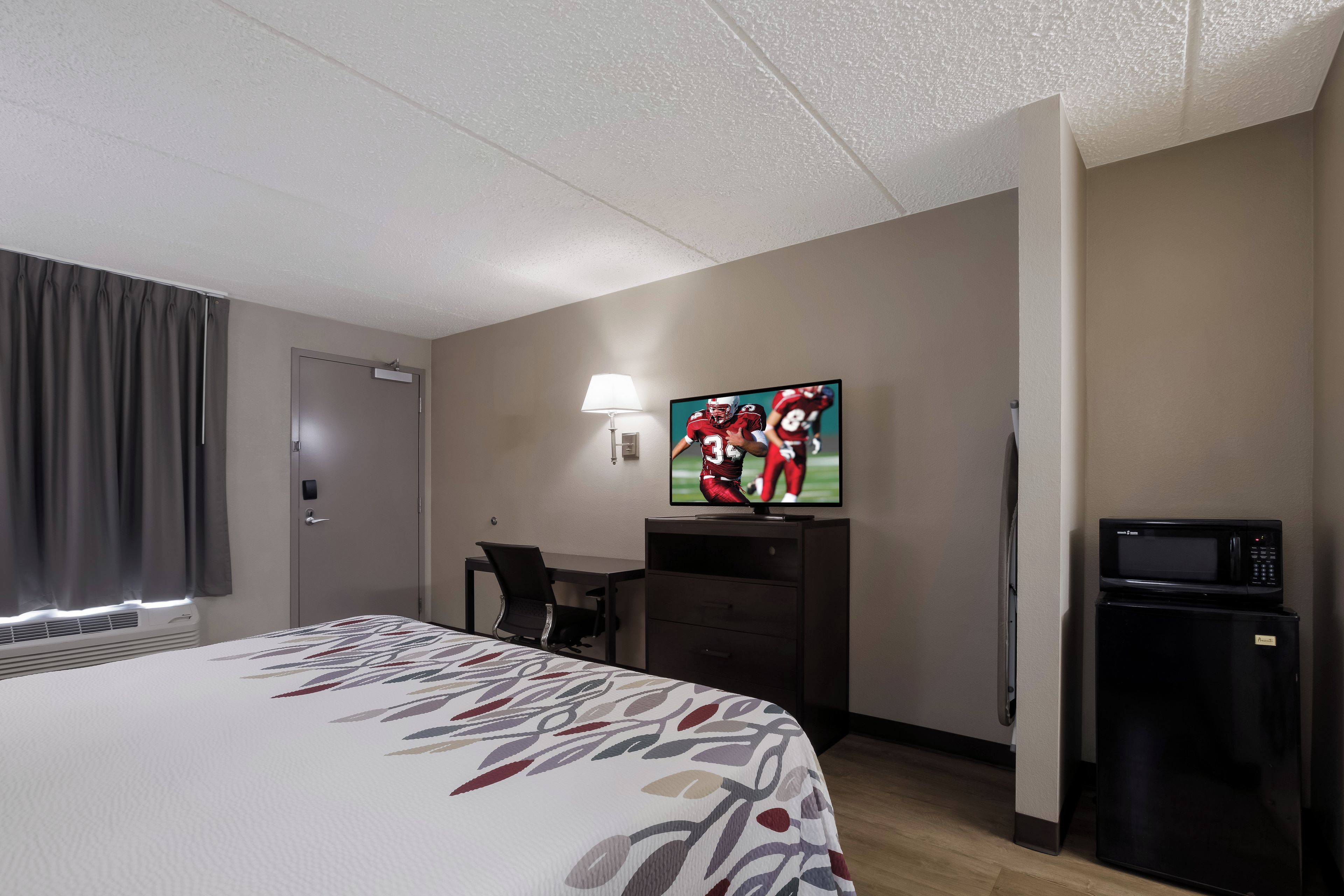 Una habitación de hotel con cama, escritorio, televisor, lámpara y refrigerador.