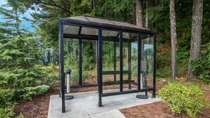 Un belvédère en verre doté d’une structure métallique, entouré d’arbres et bordé d’un chemin en béton.