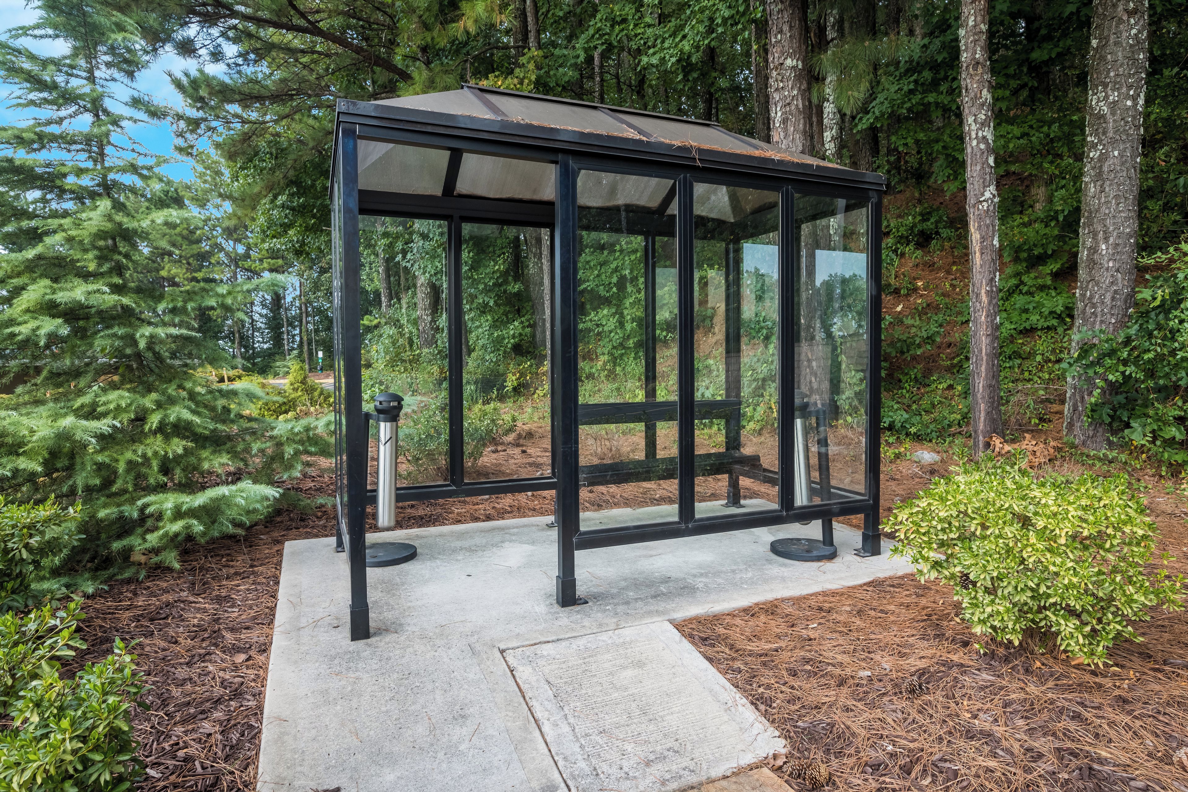 A glass gazebo with a metal frame, surrounded by trees and a concrete path.