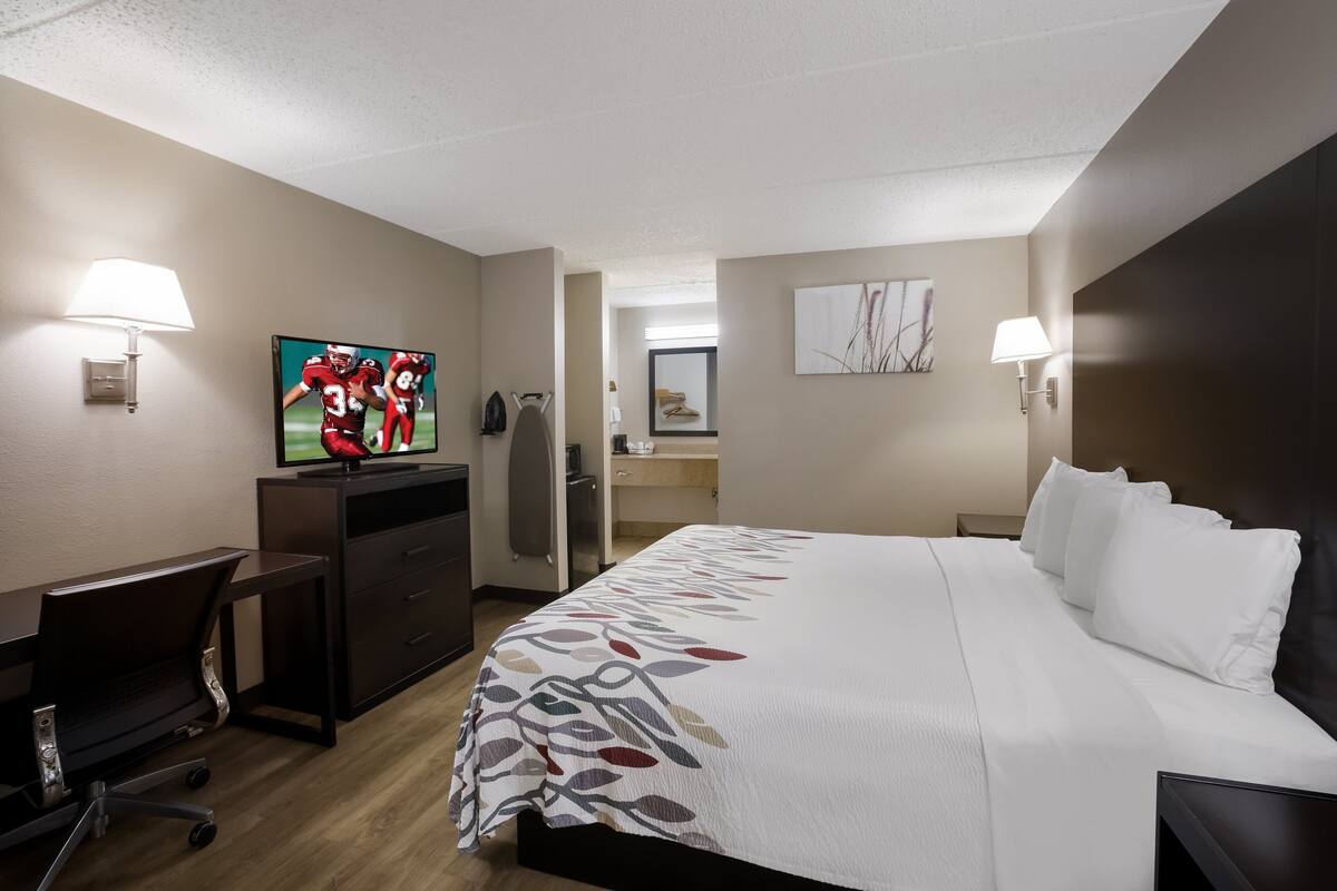 A hotel room with a bed, desk, chair, TV, and a bathroom visible in the background.