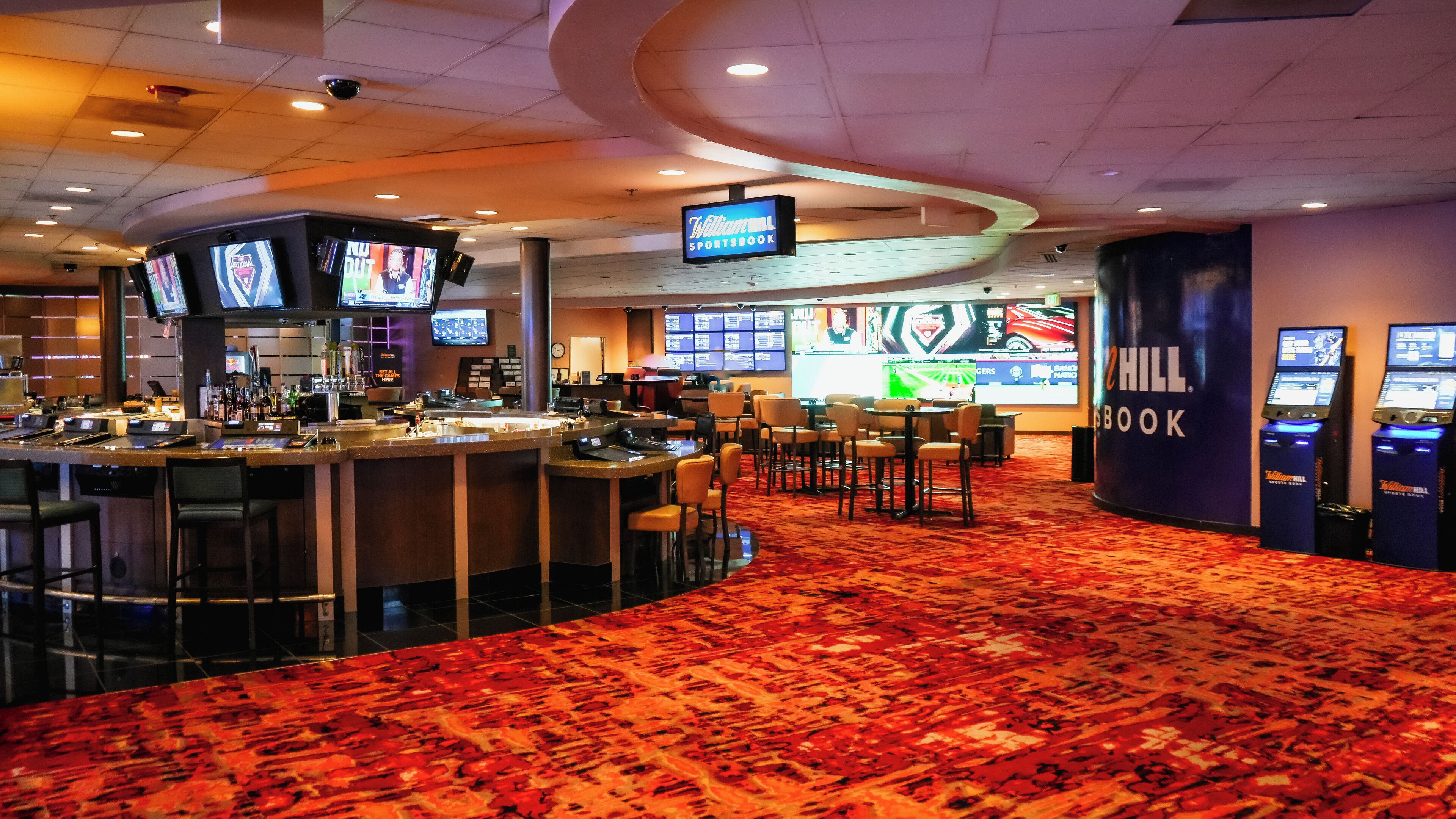 Un vestíbulo de hotel con una zona de bar, varias pantallas mostrando contenido deportivo y un gran letrero que dice 'William Hill Sportsbook'.