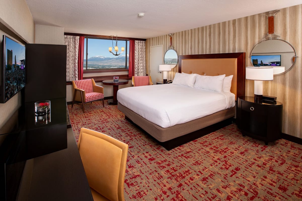 A hotel room with a large bed, a desk, a chair, a TV, and a view of mountains.