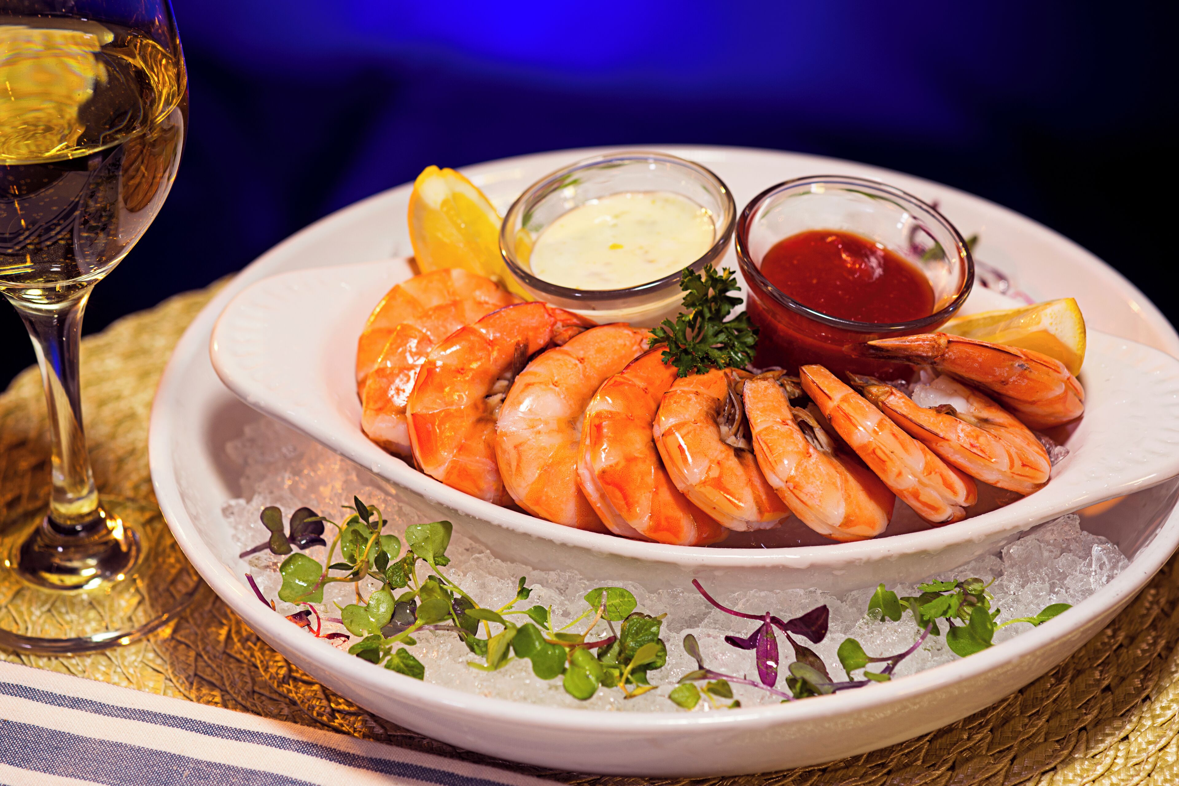 A plate of cooked shrimp with lemon wedges and two small bowls of dipping sauces, served on a bed of ice.