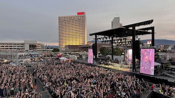 A large outdoor concert with a crowd, stage, and city buildings in the background.
