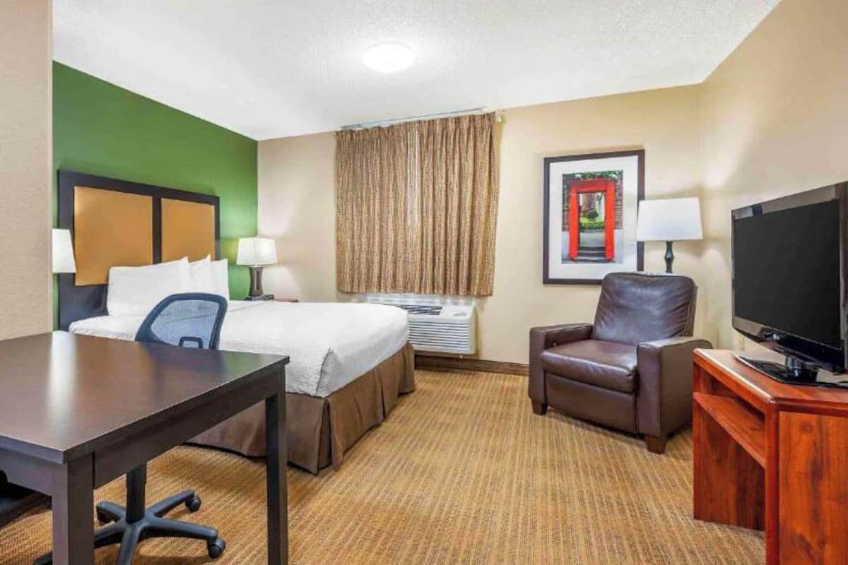 Una habitación de hotel con cama, escritorio, silla, televisión y un cuadro en la pared.