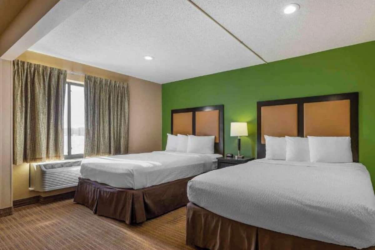 Habitación de hotel con dos camas, una ventana con cortinas, una mesita de noche con lámpara y una pared de acento verde.