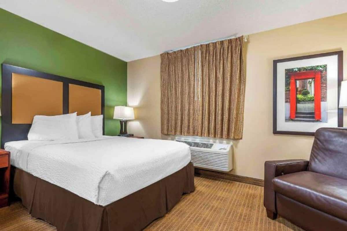 Habitación de hotel con cama, una silla, mesita de noche, lámpara, ventana con cortinas y un cuadro en la pared.