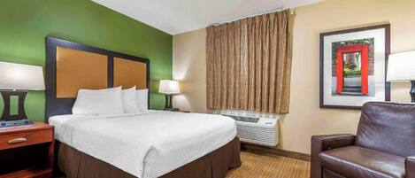 Quarto de hotel com cama, poltrona, mesa de cabeceira, luminária, janela com cortinas e um quadro na parede.