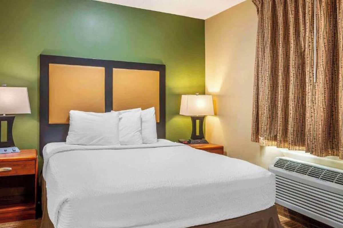 Habitación de hotel con cama, dos mesitas de noche con lámparas, una ventana con cortinas y un aire acondicionado instalado en la pared.