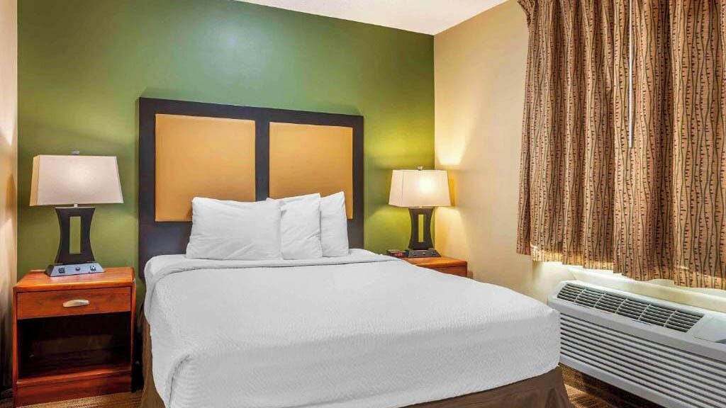 A hotel room with a bed, two bedside tables with lamps, a window with curtains, and a wall-mounted air conditioner.