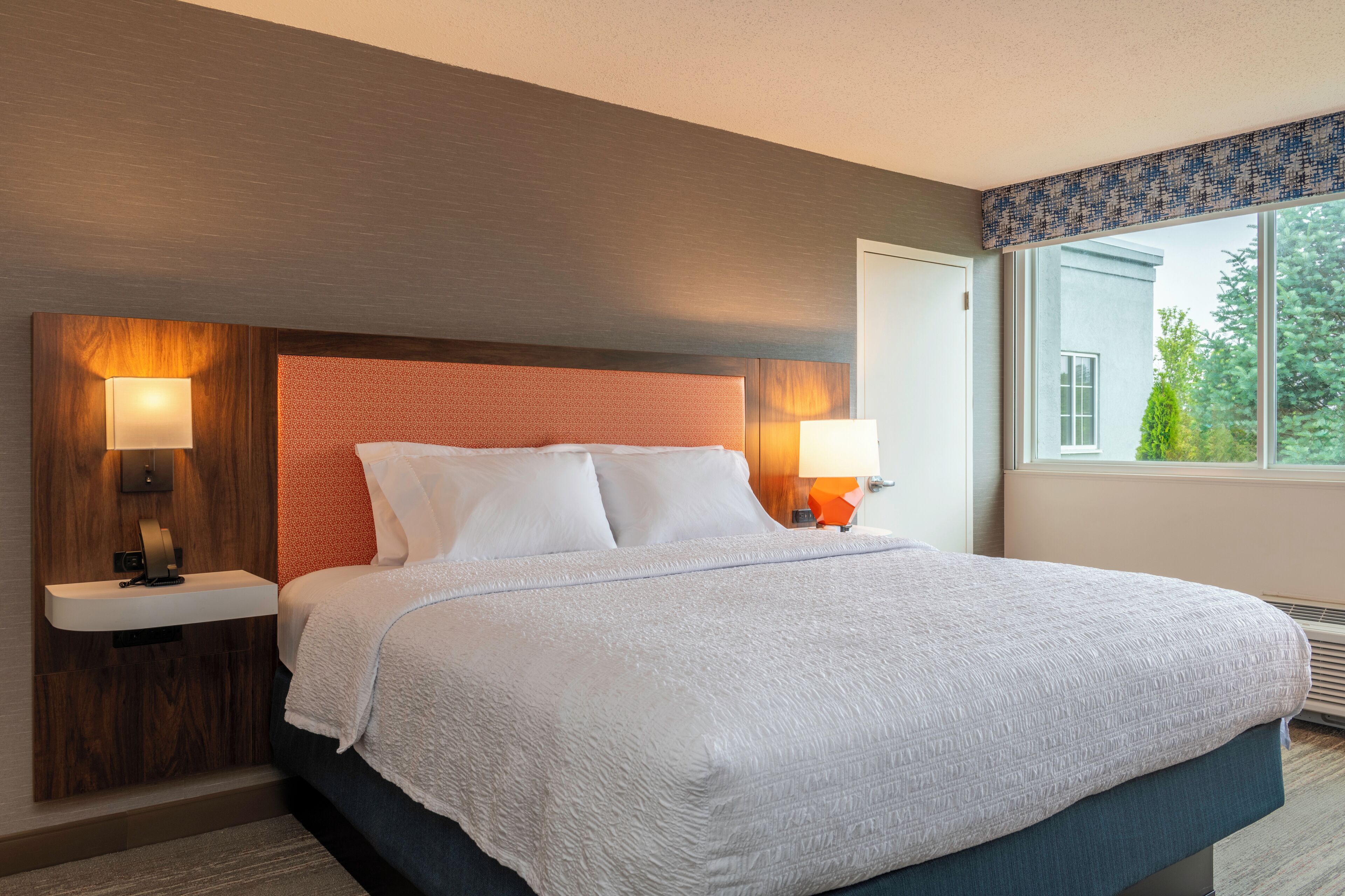 A modern hotel room with a large bed, two bedside lamps, a nightstand, and a window with a view of greenery.