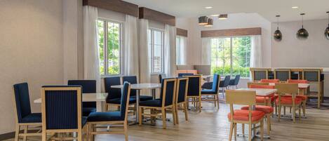 A spacious dining area with multiple tables and chairs, large windows with curtains, and a wooden floor.