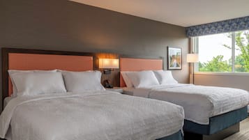 Quarto de hotel com duas camas, uma mesa de cabeceira, uma luminária e uma janela com vista para o verde.