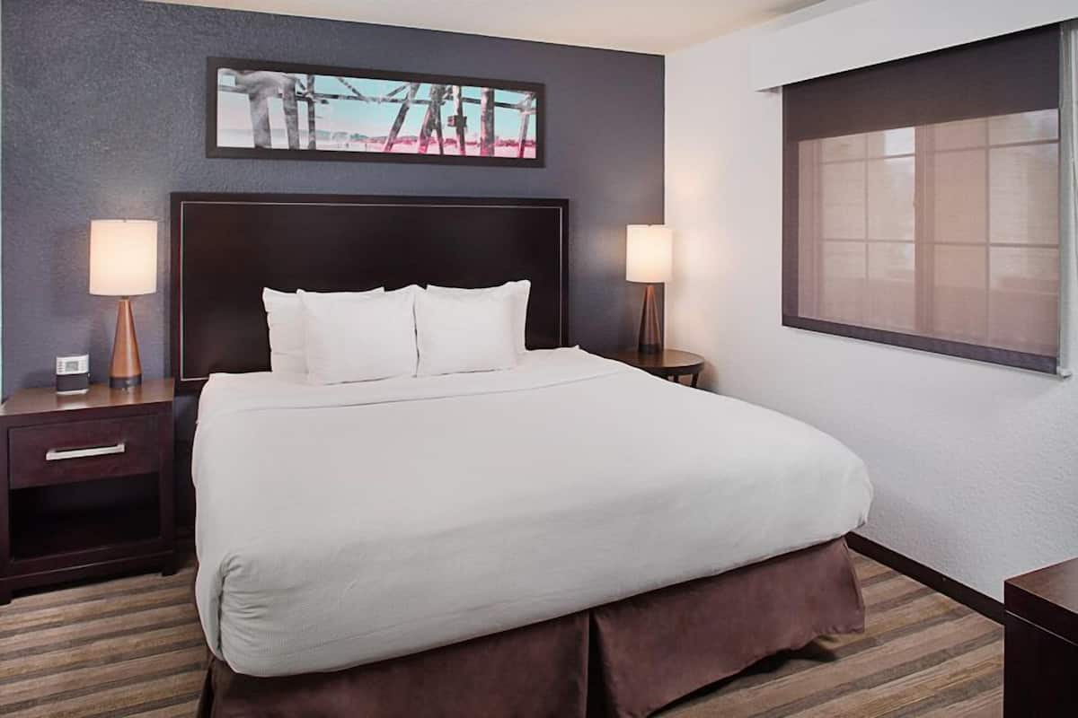 A hotel room with a large bed, two bedside tables, a nightstand, a window with blinds, and a painting on the wall.