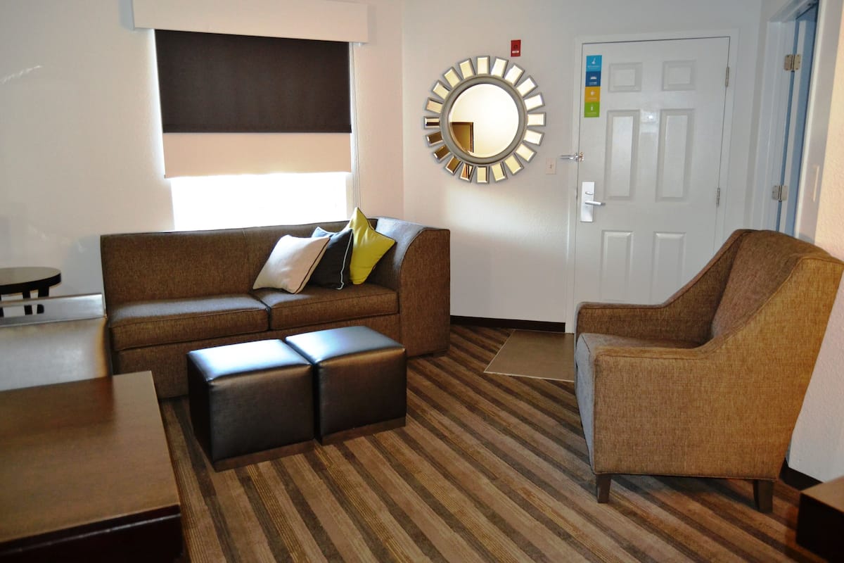 A modern living room with a brown sofa, a matching armchair, and a coffee table. A round mirror with a metallic frame is on the wall, and a door with a sign is visible.