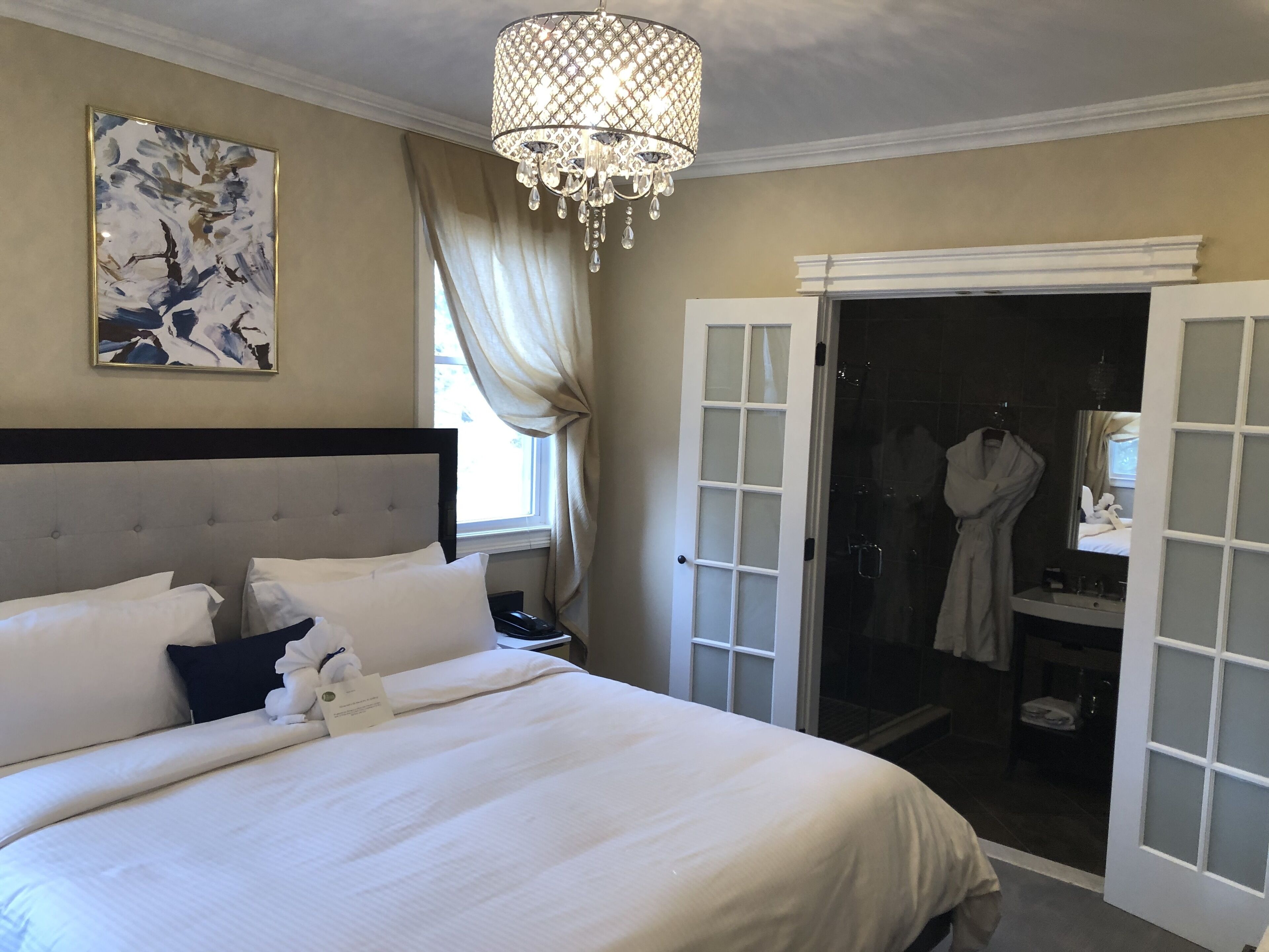Habitación de hotel con una cama grande, una araña de cristal, un cuadro y un baño visible a través de una puerta abierta.