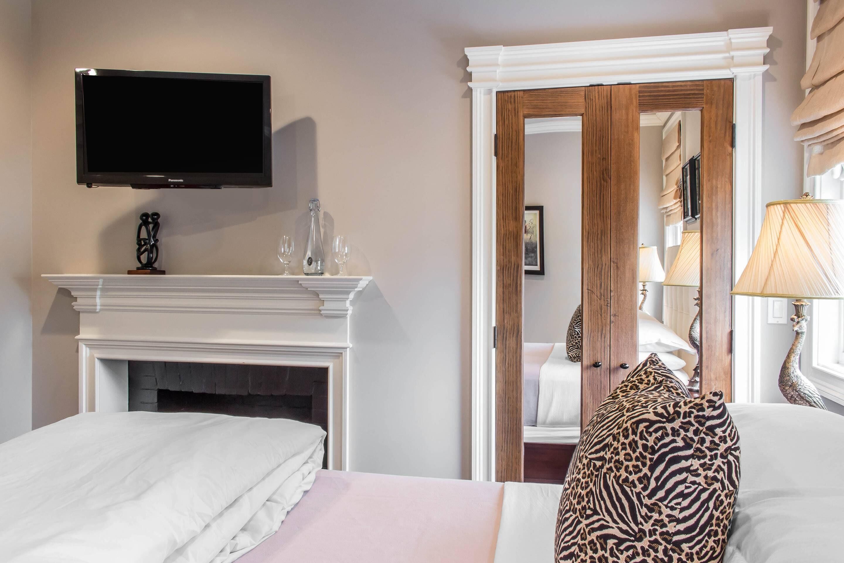 A hotel room with a flat-screen TV mounted above a fireplace, a bed with white bedding, a leopard print pillow, and a mirror reflecting the room.