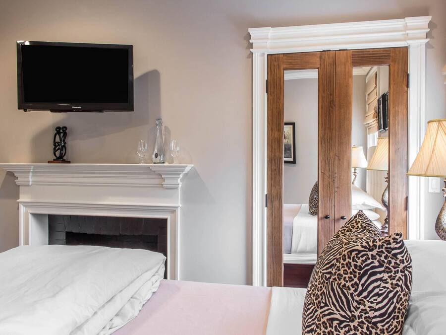 A hotel room with a flat-screen TV mounted above a fireplace, a bed with white bedding, a leopard print pillow, and a mirror reflecting the room.
