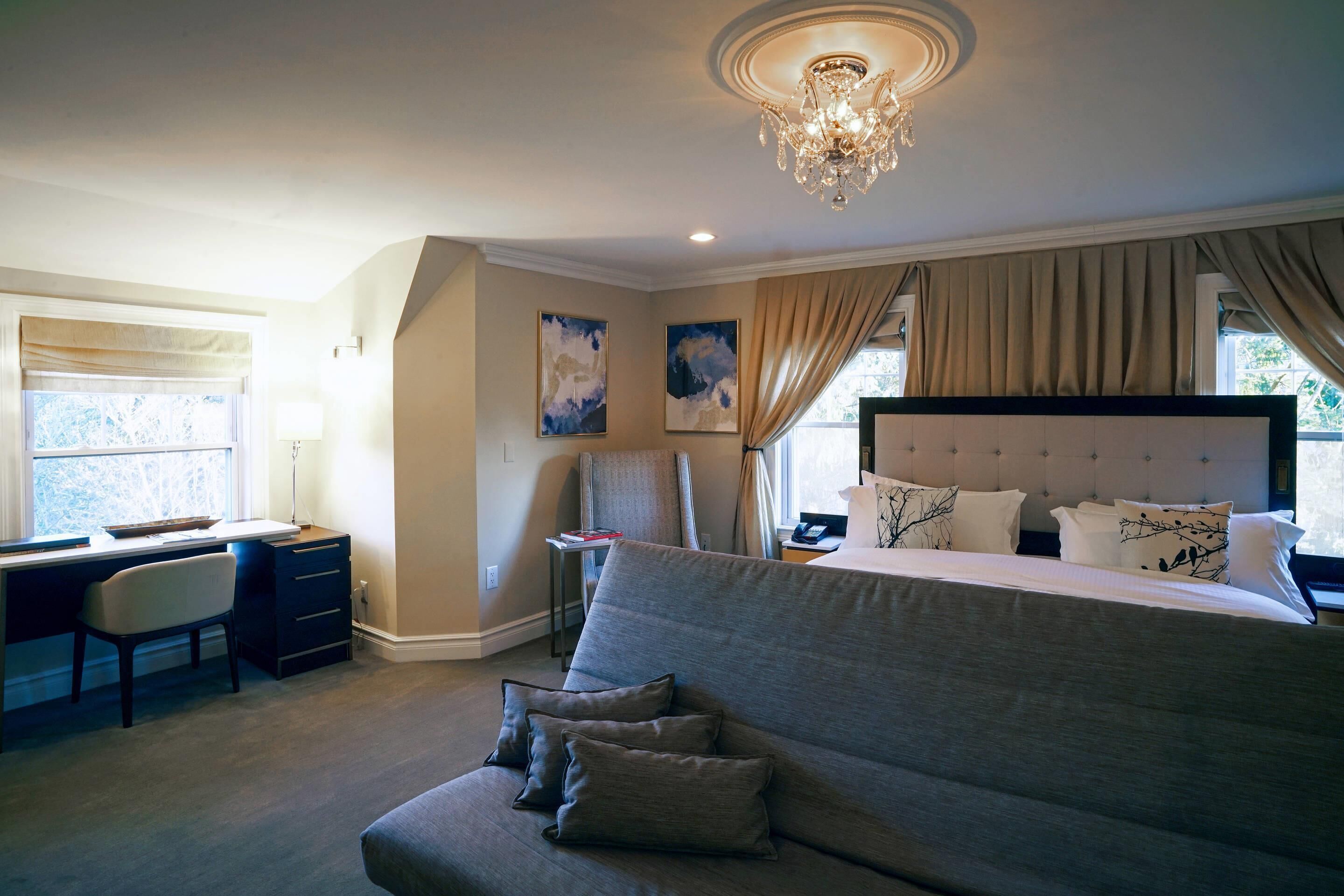A bedroom with a large bed, a desk, and a chair. There are two paintings on the wall, a chandelier, and a window with curtains.
