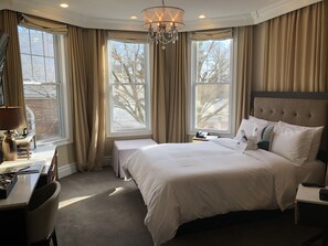 A hotel room with a large bed, a desk with a lamp, a chair, and two windows with curtains.