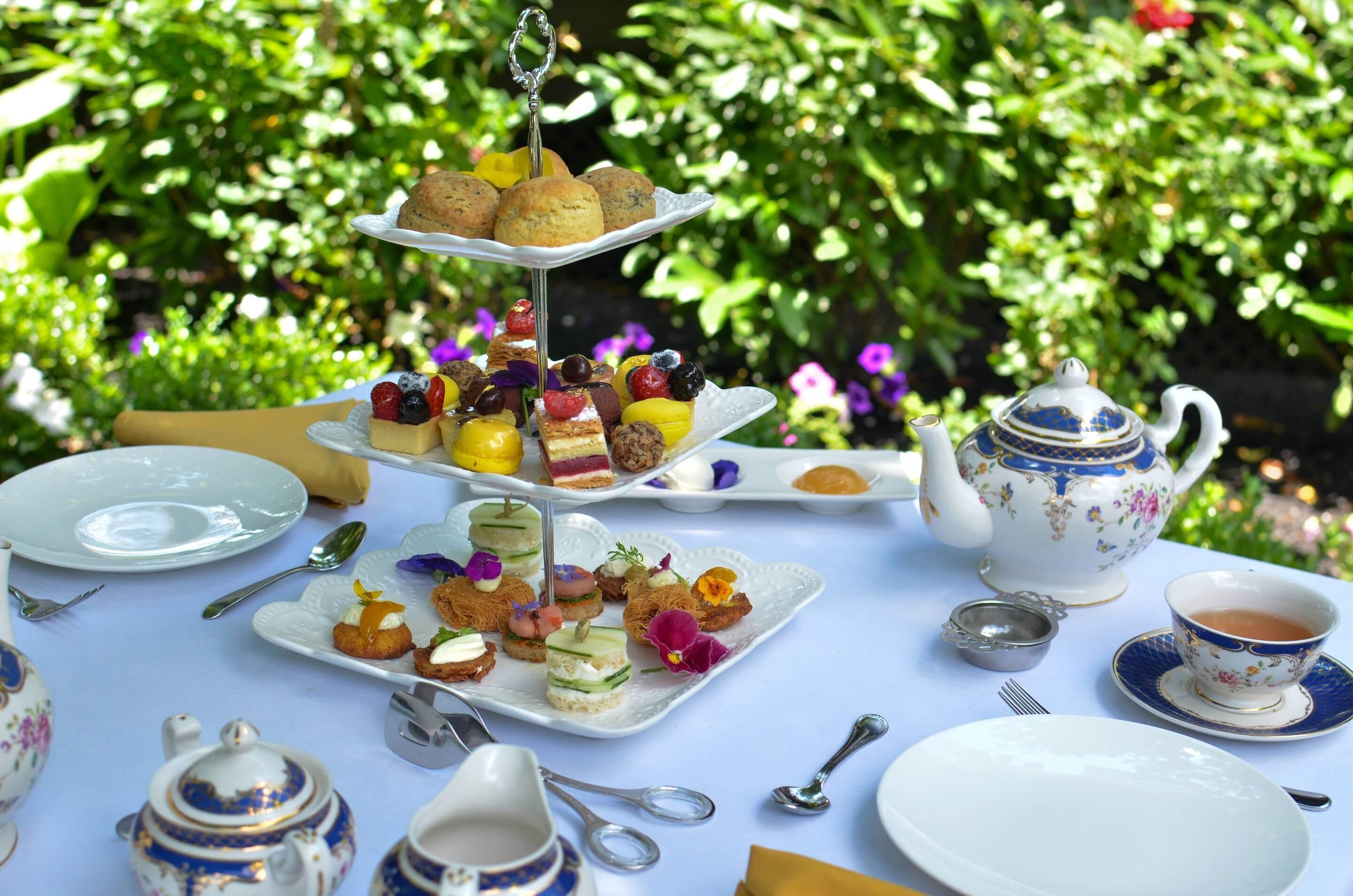 Un service à thé comprenant une théière et une tasse, un présentoir à gâteaux à plusieurs niveaux avec des pâtisseries et des fruits, et une table dressée avec des assiettes, des couverts et une serviette.