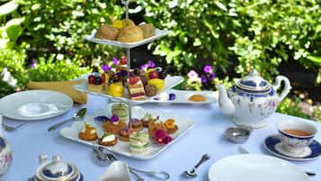 Un service à thé comprenant une théière et une tasse, un présentoir à gâteaux à plusieurs niveaux avec des pâtisseries et des fruits, et une table dressée avec des assiettes, des couverts et une serviette.