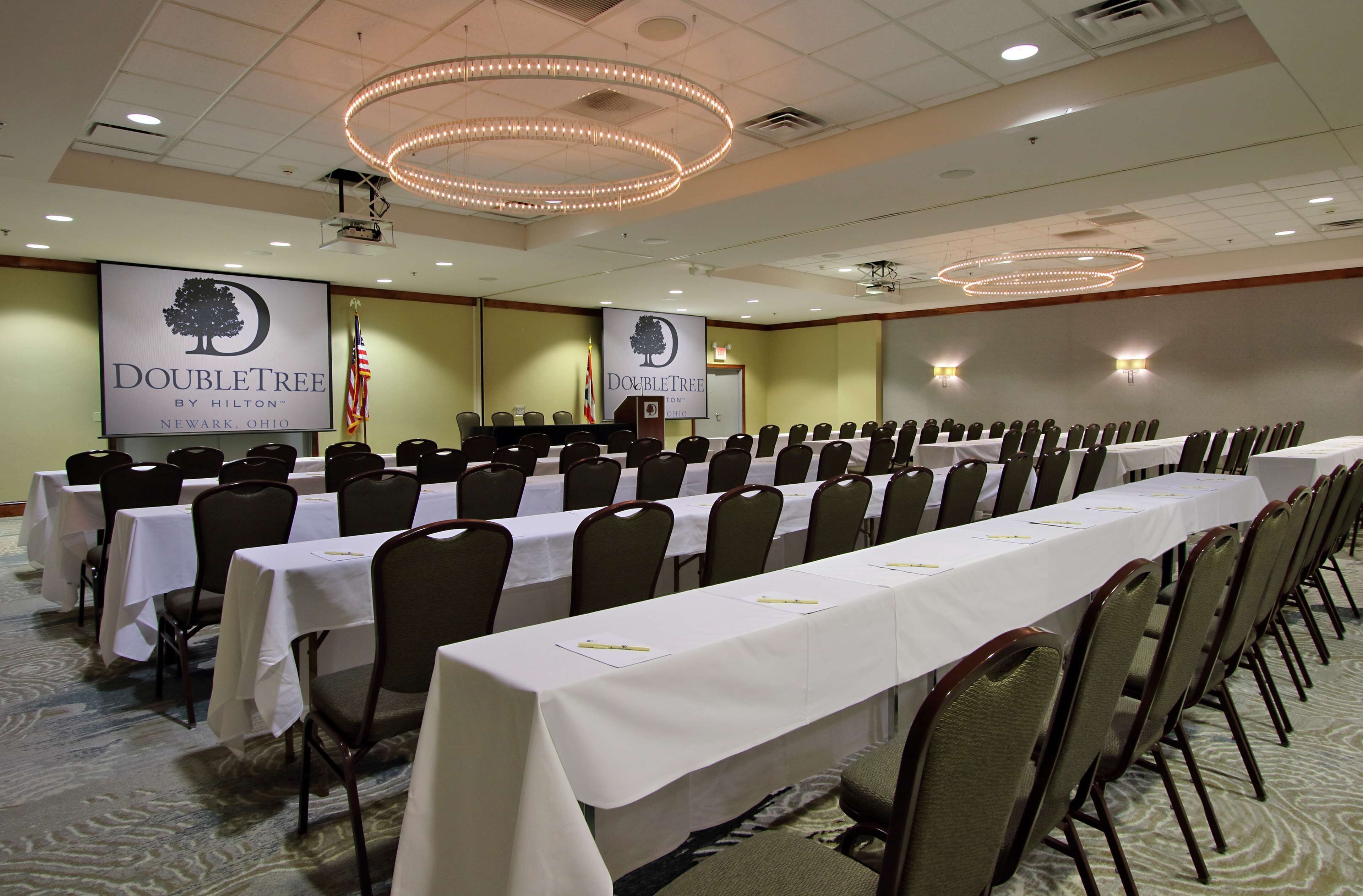 Un salón de conferencias con filas de mesas y sillas, un atril y un cartel de DoubleTree by Hilton en Newark, Ohio.