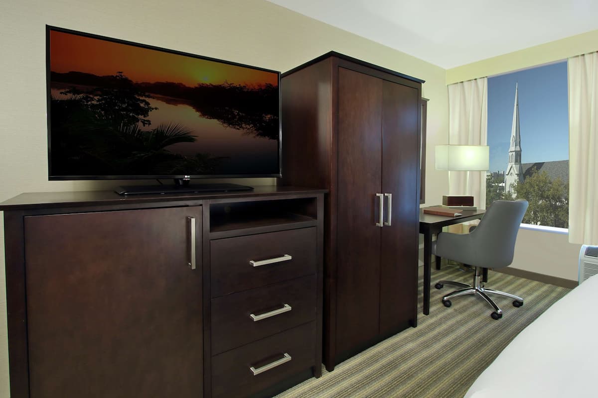 Habitación de hotel con una amplia televisión de pantalla plana, un escritorio con una silla y un ventanal con vistas a la aguja de una iglesia.