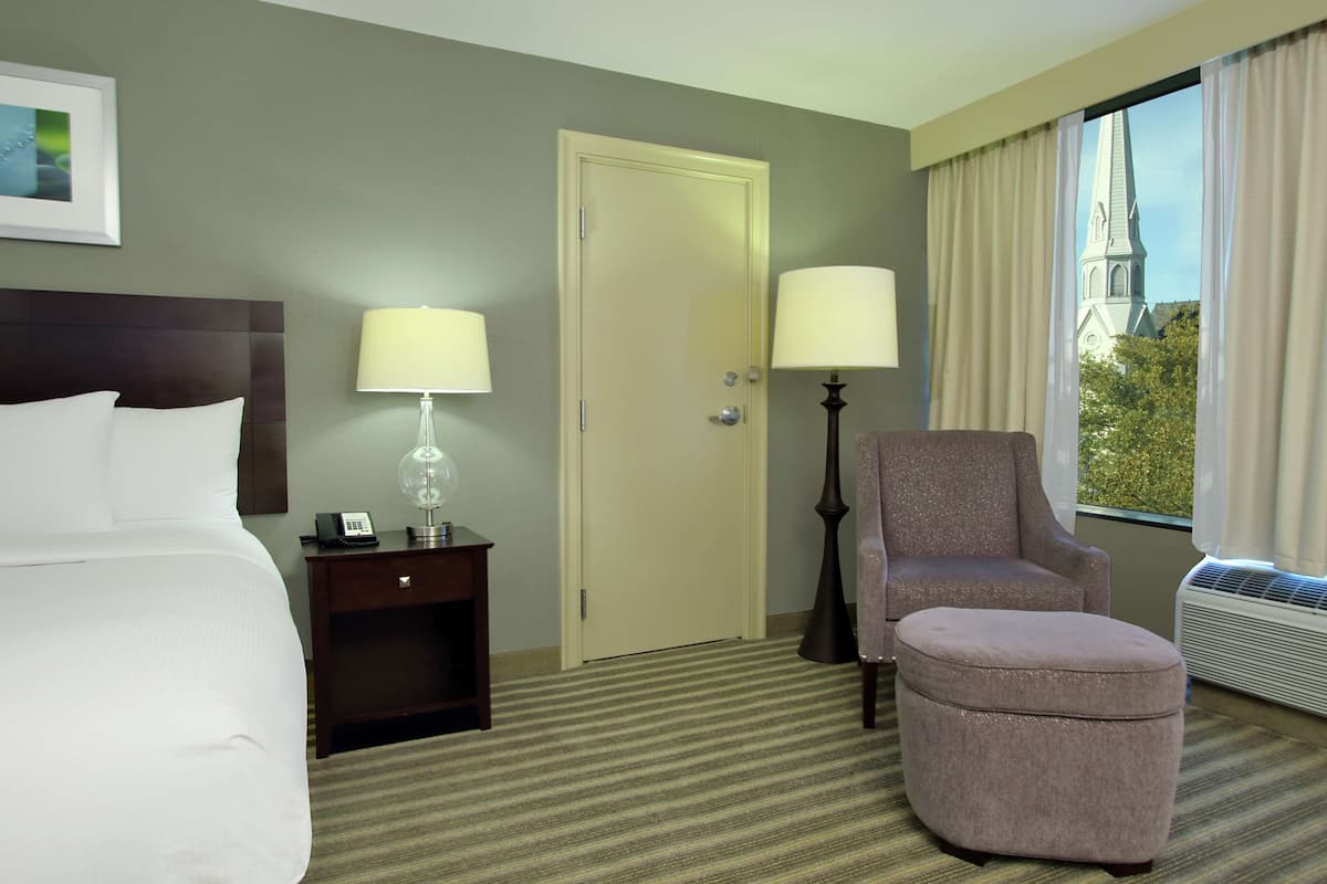 Habitación de hotel con cama, dos mesitas de noche con lámparas, una silla, un reposapiés, una puerta y una ventana con cortinas.