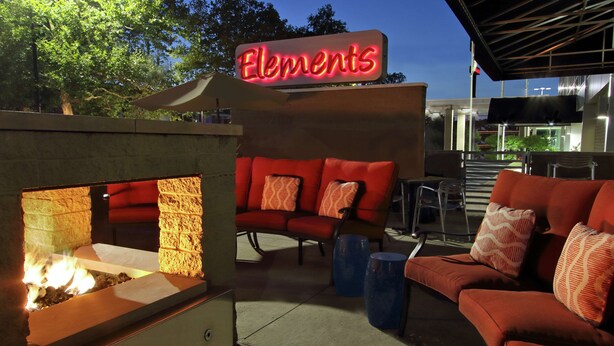 Ein Außenbereich mit Sitzgelegenheiten, einem Feuerbereich und einem Neon-Schild mit der Aufschrift 'Elements'.