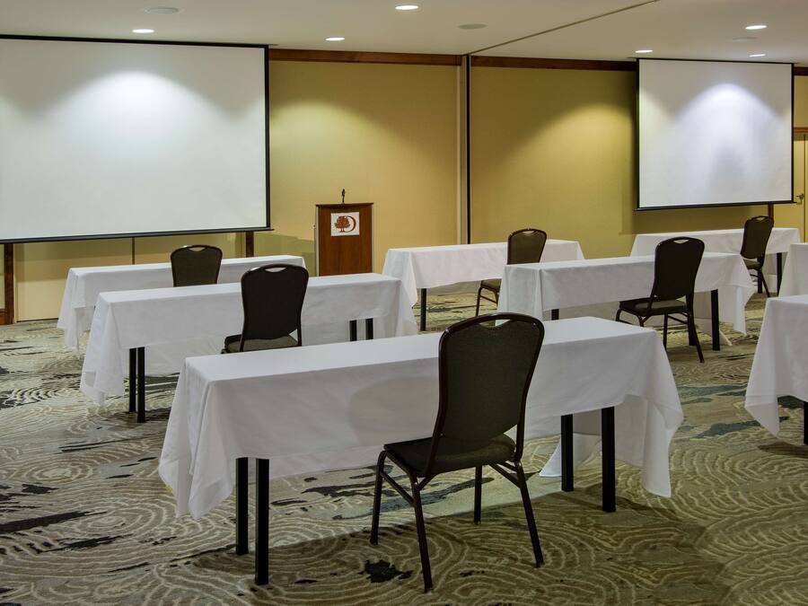 Un salón de conferencias con varias mesas y sillas, un atril y dos pantallas de proyección.