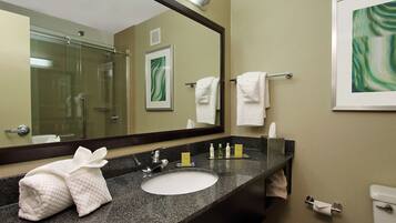A hotel bathroom with a granite countertop sink, a large mirror, a glass shower enclosure, and a framed artwork on the wall.