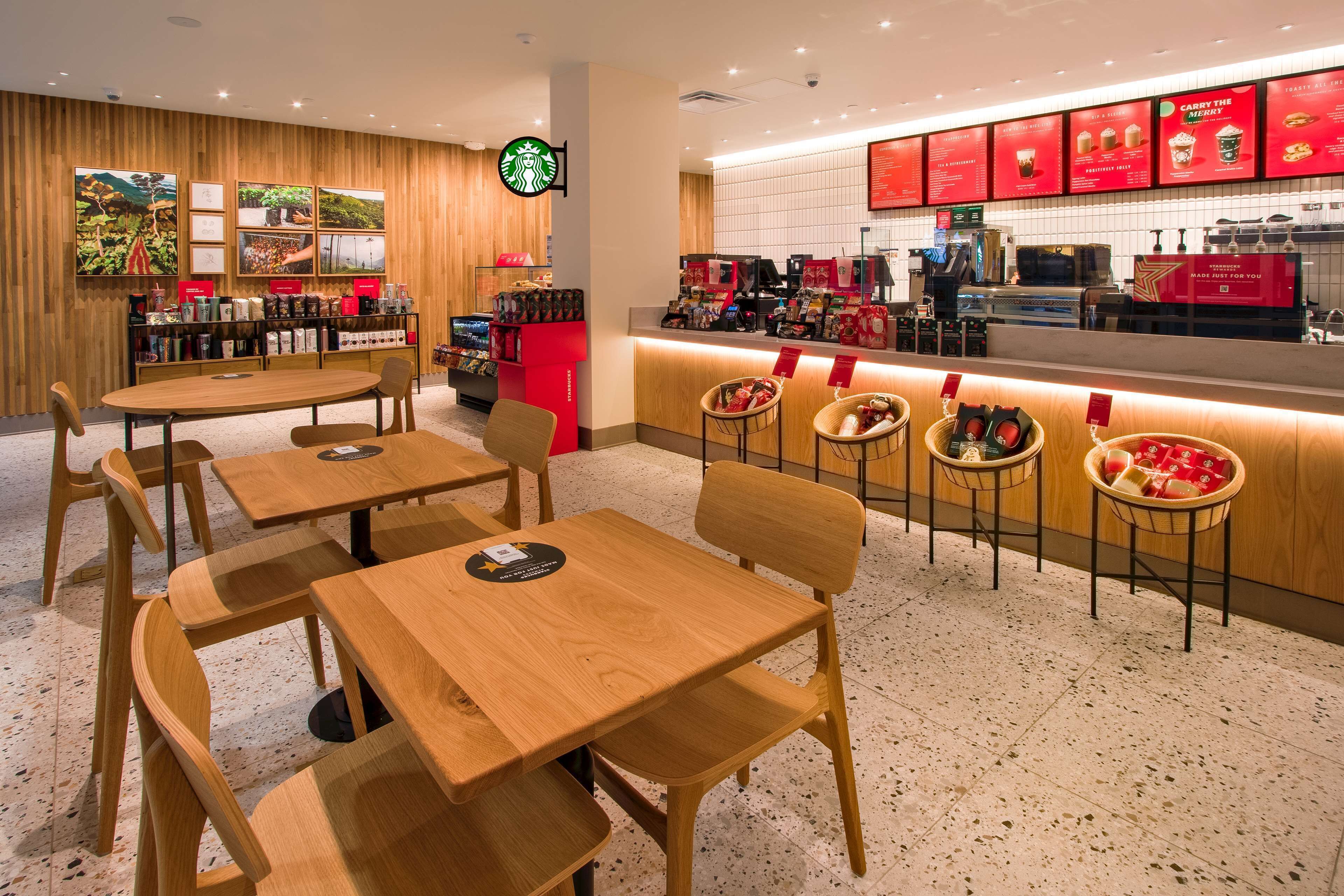 Interior de una cafetería Starbucks con mesas y sillas de madera, una barra con carteles de menú y una pared con cuadros.