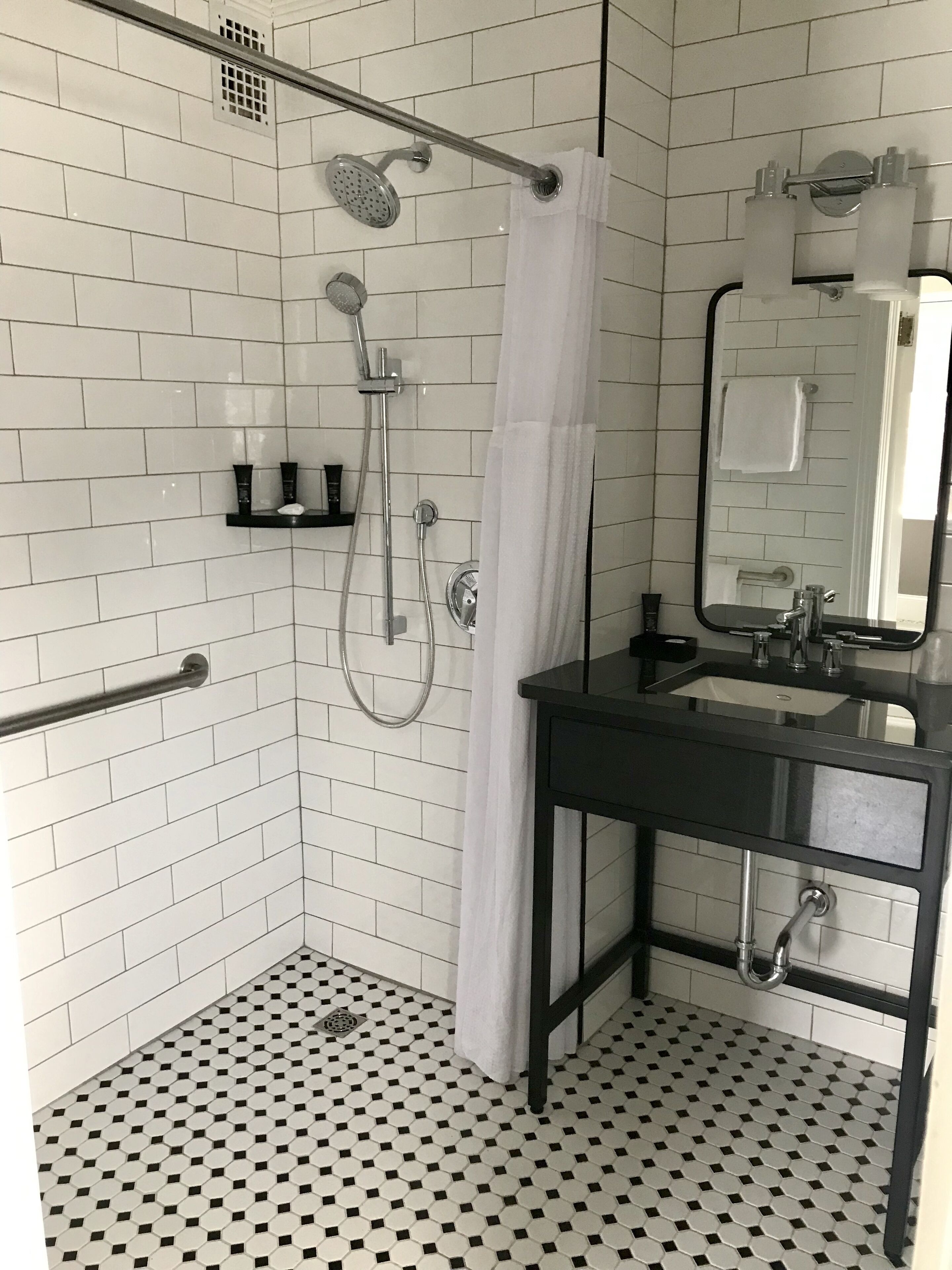 A bathroom with white subway tiles, a black and white checkered floor, a shower with a handheld showerhead, a glass-enclosed sink, and a mirror with a towel hanging on the wall.