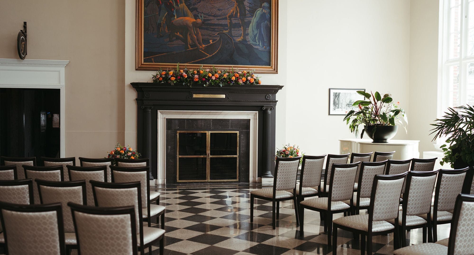 A hotel lobby with a checkered floor, a fireplace with a plaque, a large painting, and a seating area with chairs.