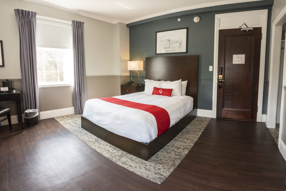 A hotel room with a large bed, a wooden headboard, a nightstand, a lamp, a window with curtains, and a door.